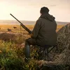 A hunter sits on a stool in a field at sunrise, holding a shotgun. Produktbild-Vorschau