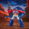 A Transformers toy, Optimus Prime, is posed in a dynamic stance holding a weapon, with a backdrop suggesting a rocky, mountainous, and possibly alien landscape. Anteprima dell’immagine prodotto