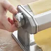 A person's hand is adjusting a knob on a stainless steel pasta maker, with a sheet of yellow dough being fed through it. Flour is visible on the wooden surface. Produktbild-Vorschau