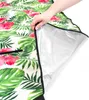 A hand is shown lifting a corner of a picnic blanket with a tropical floral pattern, revealing its silver, waterproof underside. Náhled obrázku produktu