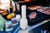 A close-up of a white FinaMill grinder on a grill counter, with ingredients and a steak on the grill in the background. Náhled obrázku produktu