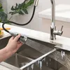 A hand is holding a pull-down faucet spray head over a stainless steel sink, with water spraying out. Podgląd zdjęcia produktu
