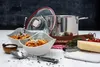 A stainless steel pot set with a glass lid is displayed on a white table, alongside two bowls of pasta, a block of cheese, tomatoes, and herbs. Anteprima dell’immagine prodotto