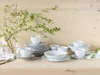 A collection of white porcelain dinnerware, including plates, bowls, and cups, arranged on a wooden surface with a vase of green branches. Náhľad obrázku produktu