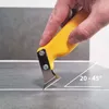 A hand is using a yellow tool to scrape a line on a tiled surface. Produktbild-Vorschau