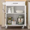 A white kitchen cart with a wooden top, open shelves, and drawers, displaying kitchenware. Podgląd zdjęcia produktu