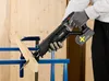 A person wearing gloves is using a cordless reciprocating saw to cut a piece of wood, which is supported by a blue stand. Produktbild-Vorschau