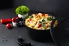A black pot filled with food, placed on a dark surface with some vegetables and a small plant nearby. Produktbild-Vorschau