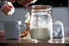 A close-up shot of a Kilner jar filled with a white powder, placed on a wooden table next to a blue measuring cup. In the background, a person's hands are visible, suggesting preparation for baking or cooking. Produktbild-Vorschau