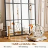 A mirror with a black metal frame is shown in a room setting, reflecting curtains and decorative items like vases and a candle holder. Produktbild-Vorschau