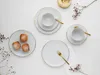 A flat lay of white porcelain dinnerware with gold trim, including plates, cups, and saucers, alongside three small pastries dusted with powdered sugar. Náhľad obrázku produktu
