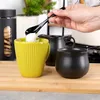 A hand is adding sugar to a yellow mug, with a black milk jug and sugar bowl nearby. Produktbild-Vorschau