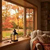 A cozy indoor scene with a bay window overlooking an autumn landscape. A black metal lantern with lit candles sits on the windowsill, next to a cup and saucer. A chair draped with warm blankets and pillows is visible on the right. Náhled obrázku produktu