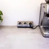 A triple power strip with a black plug is shown on a light gray countertop, with a coffee machine to the right and a plant to the left. Náhled obrázku produktu