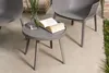 A small round table with an open book and a vase of greenery on it, placed on a patio next to a gray chair. Produktbild-Vorschau