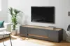 A modern living room with a TV mounted on the wall above a low media console. A large potted plant is to the left of the console, and a small side table with a glass of water is in the foreground. Produktbild-Vorschau