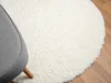 A close-up view of a round, white, shaggy rug on a wooden floor, with the armrest of a gray chair visible on the left. Náhľad obrázku produktu