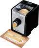 A pasta machine is shown with fresh pasta being extruded onto a wooden cutting board. The machine is black and silver, with a clear lid showing dough inside. Freshly made spaghetti is visible coming out of the front of the machine and lying on the board. Produktbild-Vorschau