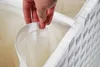 A hand is shown holding the edge of a white fabric liner inside a white woven basket, highlighting the interior of the product. Podgląd zdjęcia produktu