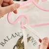 A close-up shot of a person's hands holding a pink clothes hanger with a white strap attached to it. The background is a blurry white garment with text. Produktbild-Vorschau