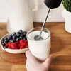 A hand is holding a glass of milk while a frother is being used to mix it. A bowl of berries is next to the glass. Produktbild-Vorschau