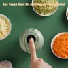A finger presses the button on a small, green electric food chopper, surrounded by bowls of chopped ingredients like garlic, celery, and carrots. Produktbild-Vorschau