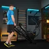 A man is standing next to a black elliptical trainer in a home gym setting. The gym has a wooden wall ladder, a chalkboard with workout notes, and blue LED strip lights. Produktbild-Vorschau