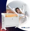 A woman is relaxing in a chair with her arms stretched up, while a BROLLUX electric heater is in the foreground. Produktbild-Vorschau