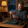 A man is sitting at a wooden table, holding up a Christmas-themed tie and looking at it with a smile. The scene is set indoors, with a fireplace and Christmas lights visible in the background, suggesting a cozy holiday atmosphere. Náhľad obrázku produktu
