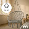 A light gray hanging chair with tassels is shown in a room with sunlight streaming through a window. A small side table with a lamp and a bowl is visible. Náhled obrázku produktu