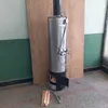A stainless steel wood-fired water heater with a shower head and faucet is standing on a floor in front of a wooden board. A small pile of firewood is on the floor next to it. Produktbild-Vorschau