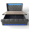 A grey upholstered bed frame with the mattress support slats raised, revealing storage space underneath. Blue LED lighting is visible along the headboard and the base of the frame. Produktbild-Vorschau