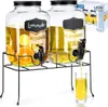 Two glass beverage dispensers with taps on a black metal stand, filled with yellow liquid and fruit slices. One dispenser has a chalkboard label that says "Lemonade". A glass is being filled from one of the taps. Náhľad obrázku produktu