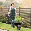 A woman is pushing a wheelbarrow with plants in a garden, behind a decorative fence. Náhled obrázku produktu