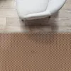 A top-down view of a rug with a geometric pattern placed on a wooden floor next to a white armchair. Podgląd zdjęcia produktu