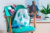 A collection of items, including a bikini, a drawstring bag with a cactus print, a blue water bottle, and a plant in a pot with a mustache, are arranged on a teal chair and a white surface. Anteprima dell’immagine prodotto