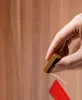 A hand is holding a brown repair wax stick over a wooden surface, applying it to a scratch. A red tool is also visible. Podgląd zdjęcia produktu