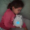 A young girl sleeping in bed, holding a plush hedgehog toy that projects lights. Voir les images du produit