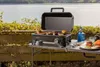 A portable grill is set up on a table outdoors next to a body of water, with food cooking on the grill. Náhľad obrázku produktu