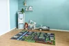 A child's play rug with a city street design is laid out on a wooden floor in a room with light blue walls. Various toys are scattered around the rug, including a toy car, a storage bin with stuffed animals, and building blocks. Produktbild-Vorschau
