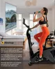 A woman is exercising on an exercise bike in a room, with a large screen displaying a scenic road. Produktbild-Vorschau