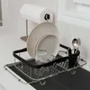A dish rack with plates, a glass, and cutlery is placed over a sink, with a paper towel holder in the background. Produktbild-Vorschau