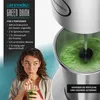 A woman drinks a green smoothie while a blender prepares another batch. Anteprima dell’immagine prodotto