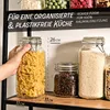 A hand is holding a large glass jar filled with pasta, placed on a shelf with other glass jars containing coffee beans and rice. Text overlays provide product information. Podgląd zdjęcia produktu
