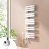A white electric towel radiator mounted on a pale pink wall next to a window with greenery visible outside. Náhled obrázku produktu