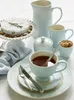 A light blue ceramic latte cup filled with tea, placed on a saucer, with a spoon inside. The cup is part of a tea set that includes a pitcher, creamer, sugar bowl, and a cake stand with a cake on it. Produktbild-Vorschau