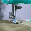 A close-up view of a sewing machine needle and presser foot stitching on a light green fabric. Produktbild-Vorschau