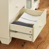 A close-up view of an open drawer in a cream-colored cabinet, showing folded white linens and a gray quilted item inside. Voir les images du produit