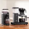 Two black coffee machines are displayed on a wooden countertop in a kitchen setting. Produktbild-Vorschau