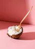 A half coconut filled with white coconut flesh, with a wooden spoon inserted into it, set against a pink background with shadows. Produktbild-Vorschau
